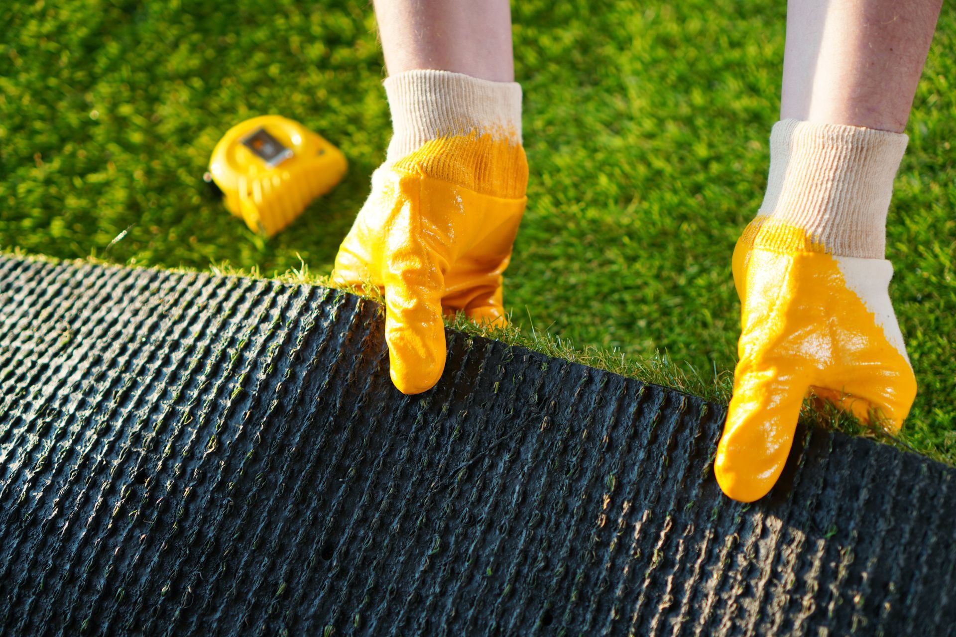 Gloved hands rolling out black turf on grass beside a yellow tape measure