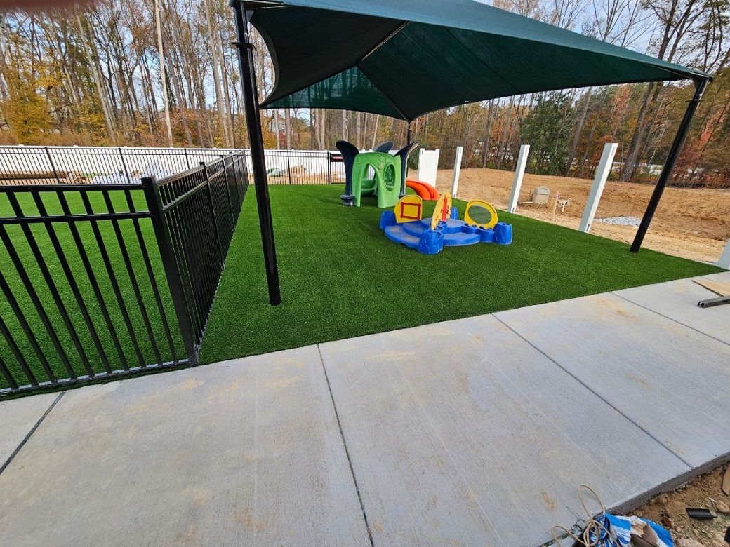 Playground under green canopy beside concrete patio and black fence, with colorful toy equipment on grass
