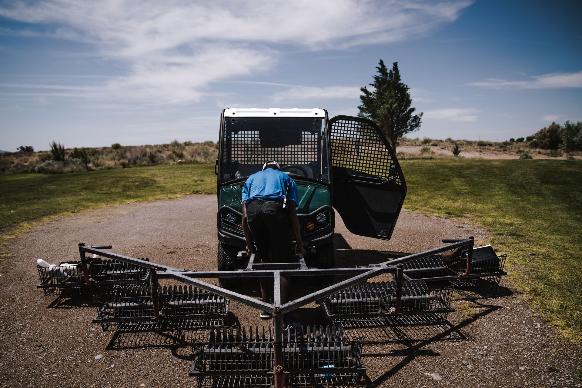 Front view of a blue tractor with a wide cultivator on a gravel path under a partly cloudy sky
