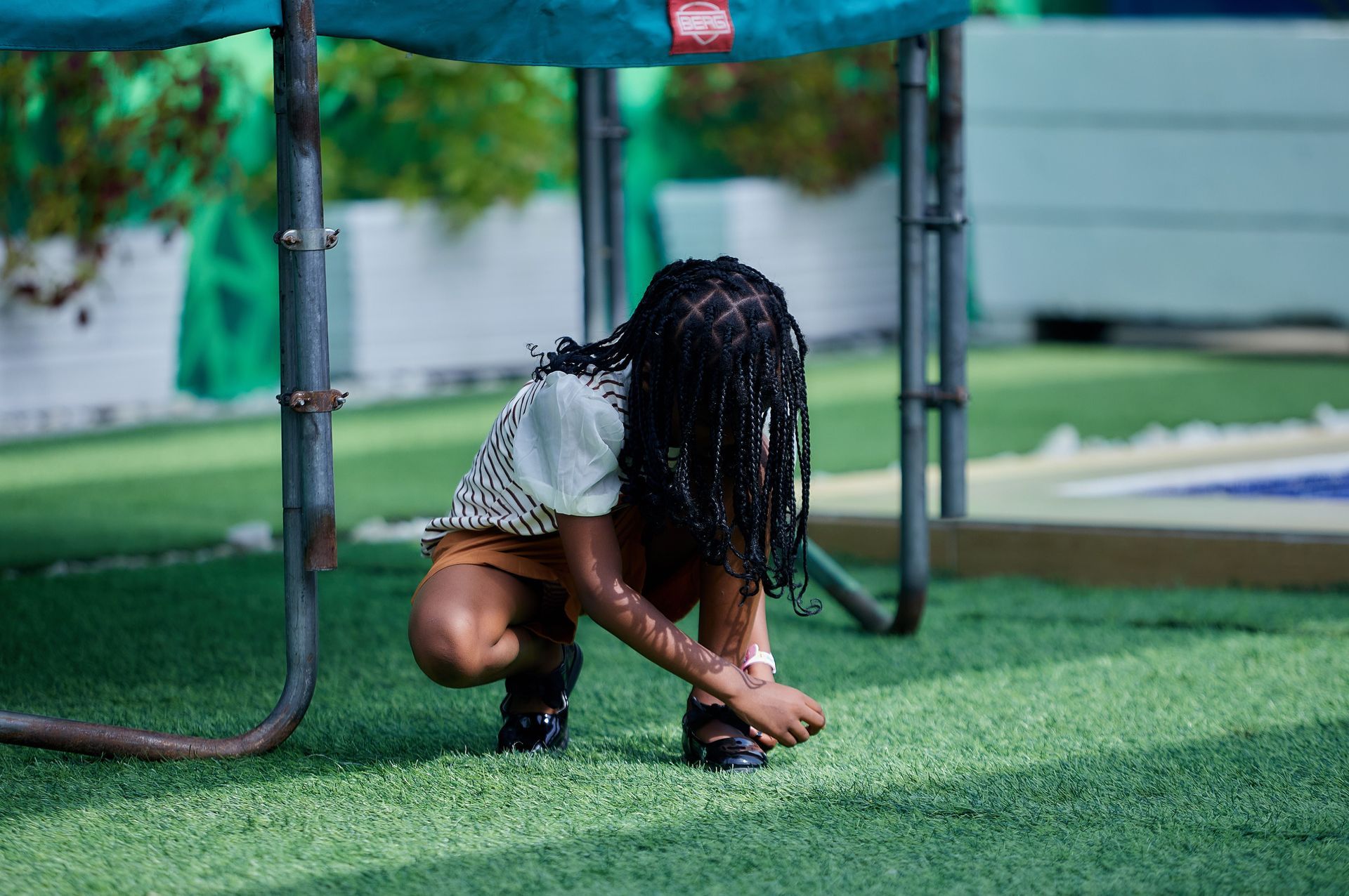 Child crouching on green turf, tying shoelaces under a metal play structure.