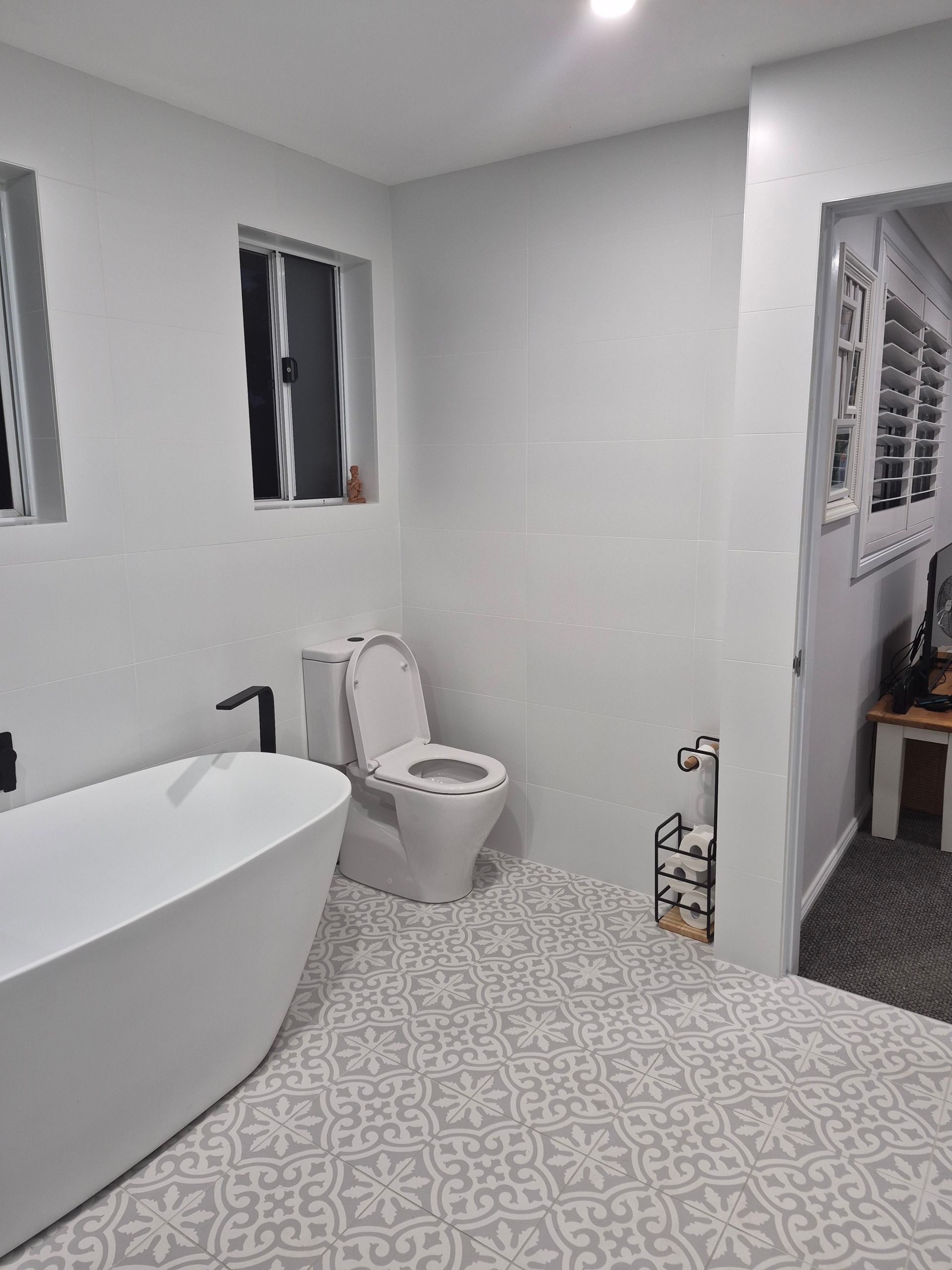 A modern bathroom featuring a white freestanding tub, a toilet, a patterned tiled floor, and two windows.