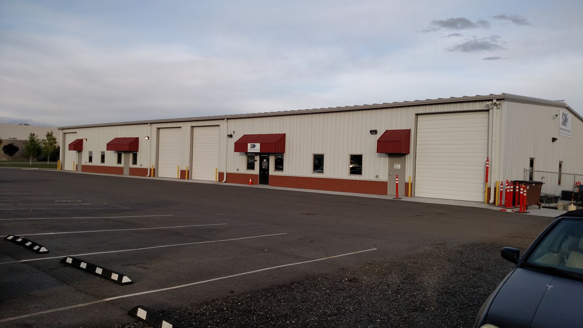 A beige warehouse building with multiple roll-up doors and red awnings, situated next to a large, empty parking lot.