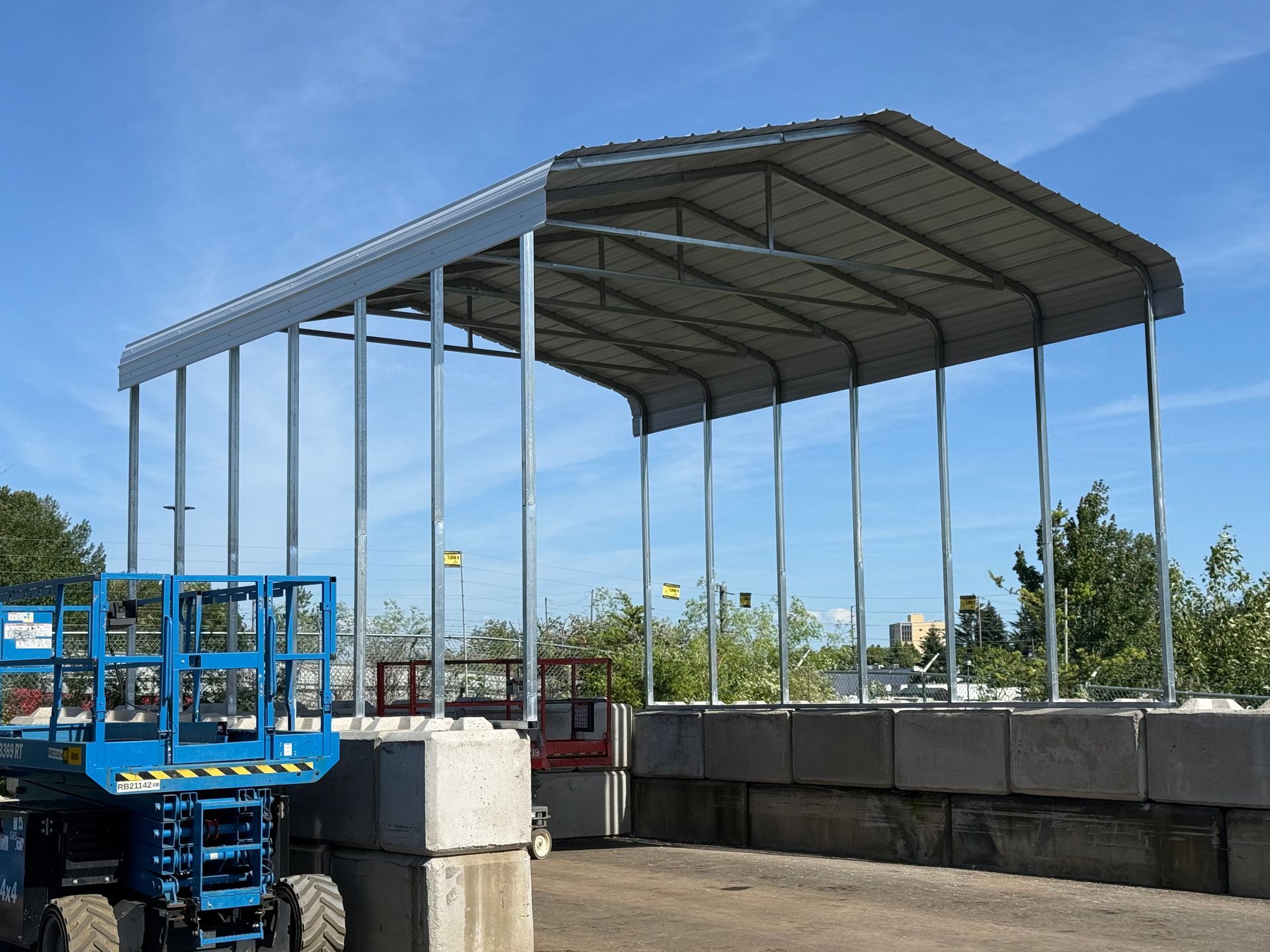Steel-roofed structure under construction beside a blue lift and concrete barriers under a clear sky
