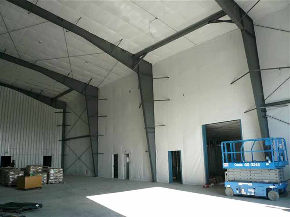 A spacious warehouse interior with high ceilings, large metal support beams, white walls, and a blue scissor lift.