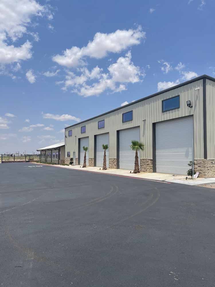 A long, metal-sided commercial building with four large garage doors, stone accents, and small palm trees under a blue sky.