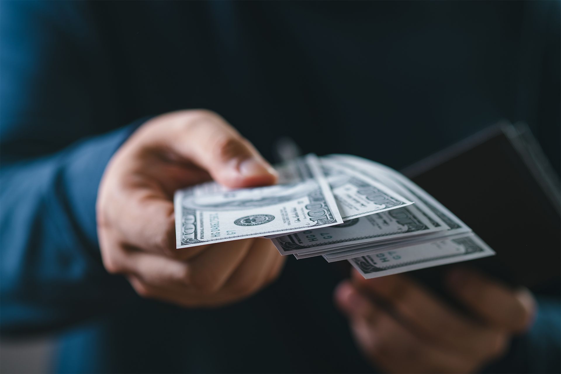 Person holding a wallet, offering a stack of U.S. hundred dollar bills, likely as payment.