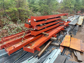A pile of orange steel I-beams stacked on wooden dunnage in an outdoor, wooded construction area.