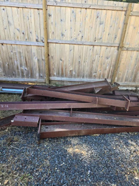 A stack of rusty brown steel I-beams lying on a gravel surface in front of a wooden fence.