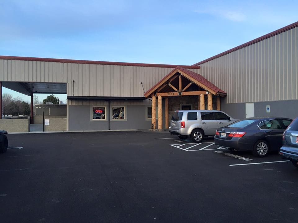 A tan, metal-sided commercial building with a wooden gabled entryway and a small parking lot with three parked cars.