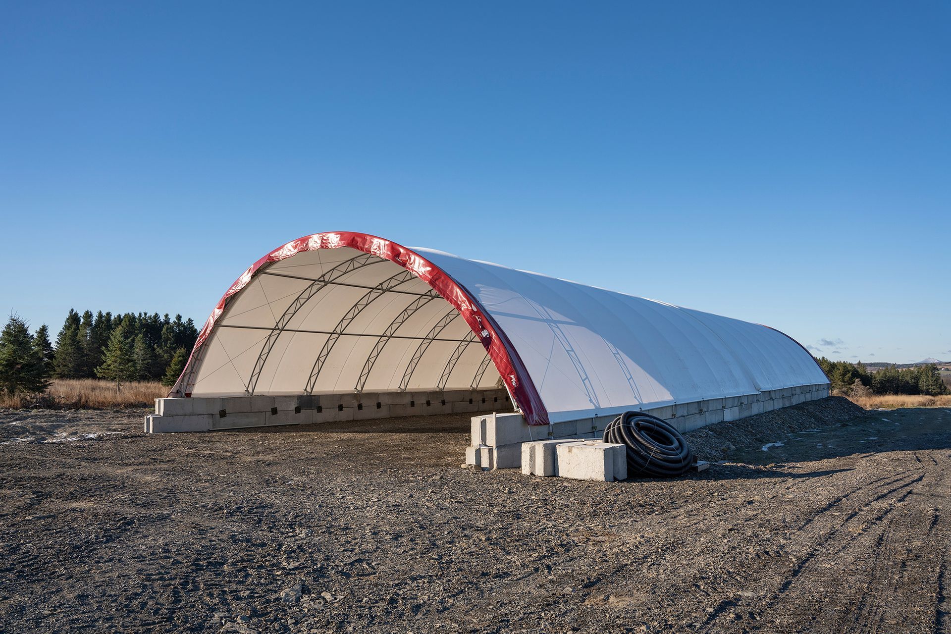 White and red arched fabric shelter on concrete blocks in a gravel field under a blue sky.