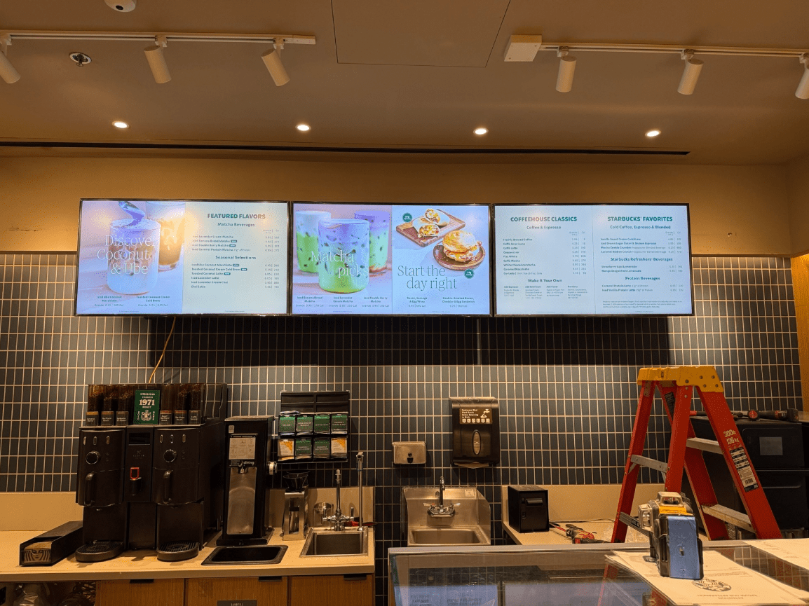Coffee shop counter with digital menu boards, espresso machines, and a red ladder by the wall