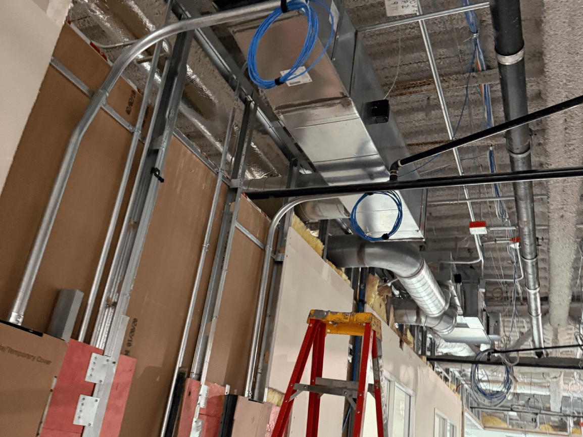Construction site ceiling with metal ductwork, exposed wiring, and a red ladder beneath unfinished drywall.