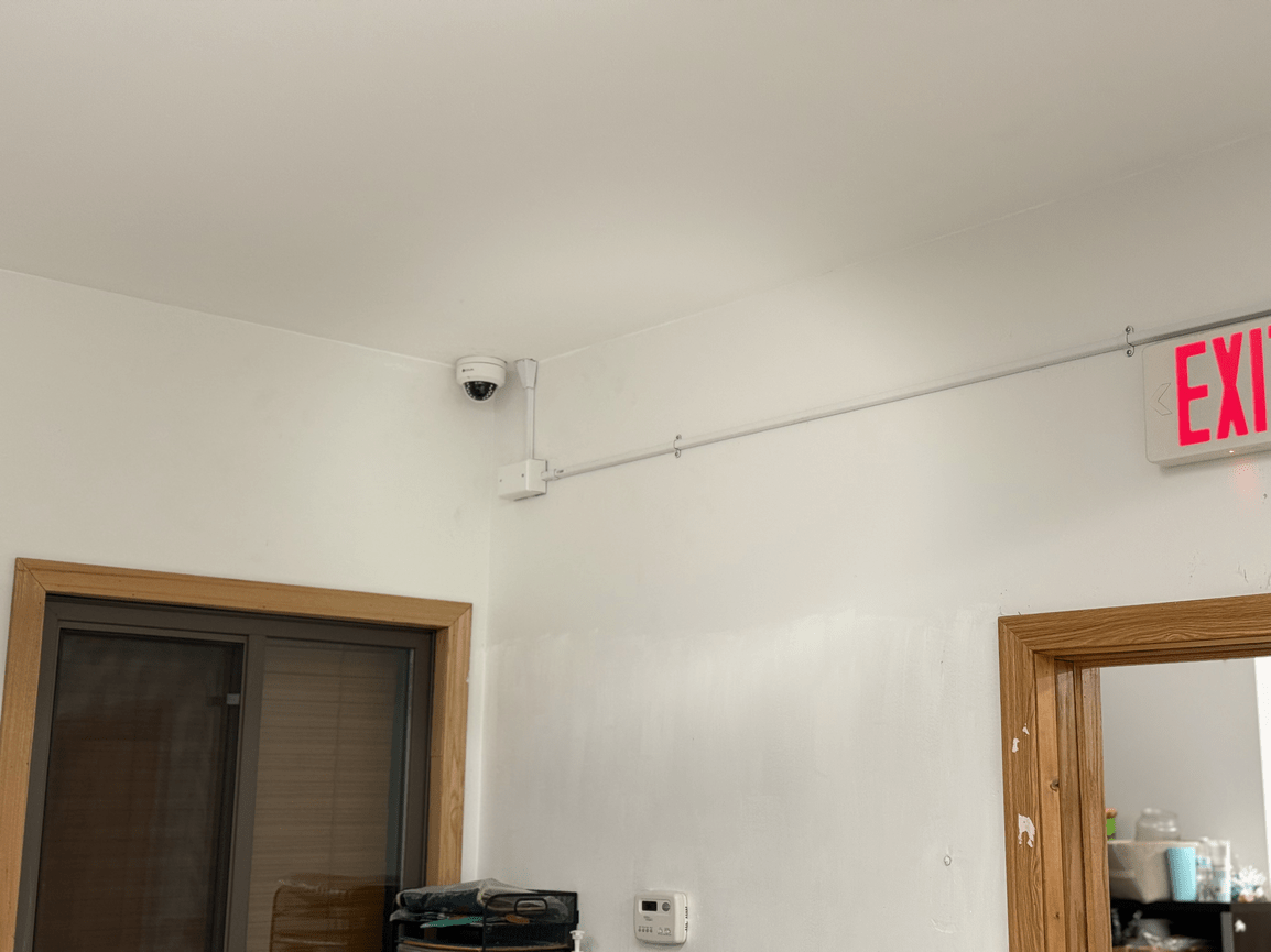 Ceiling corner with security camera, doorways, and red EXIT sign in a white hallway