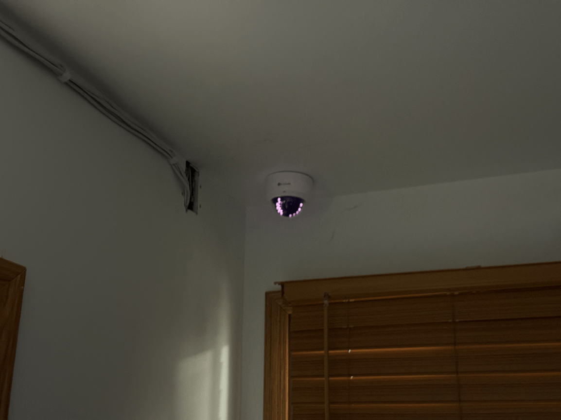 Indoor ceiling corner with a small security camera above a window with wooden blinds.