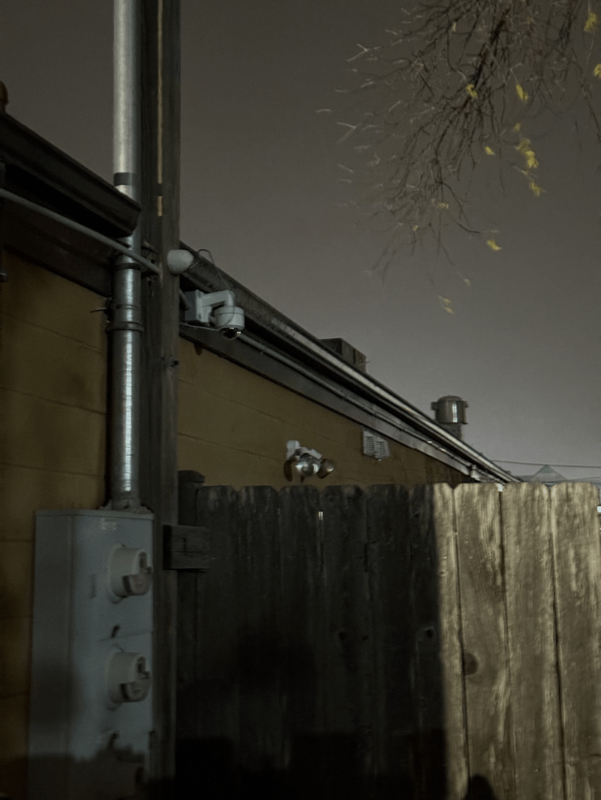 Utility pipes and electrical boxes mounted on a building wall above a wooden fence at night.