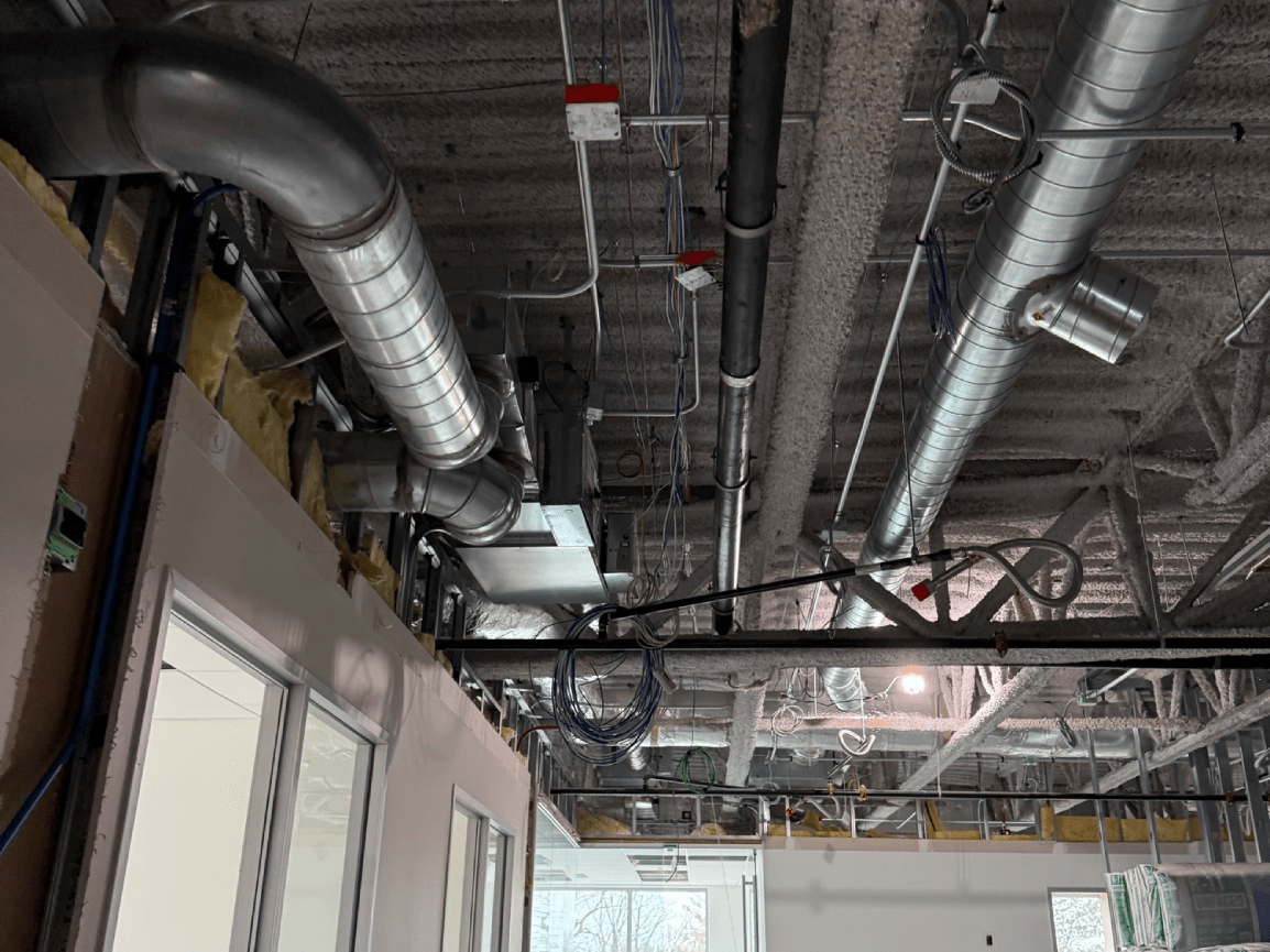Industrial ceiling with exposed ductwork, pipes, and vents above a hallway interior