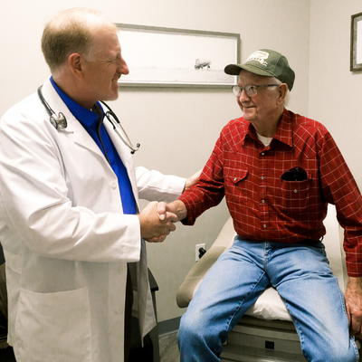 Doctor in white coat shaking hands with seated patient in red plaid shirt in exam room