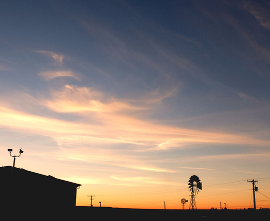 Sunset sky with orange horizon, wispy clouds, and silhouetted rooftops, poles, and trees.
