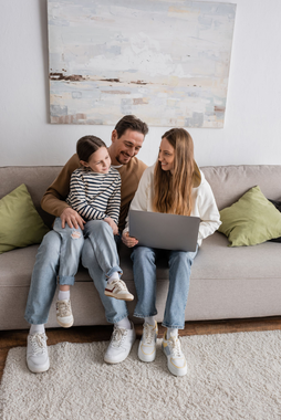 Family sitting on a couch, smiling and looking at a laptop in a bright living room.