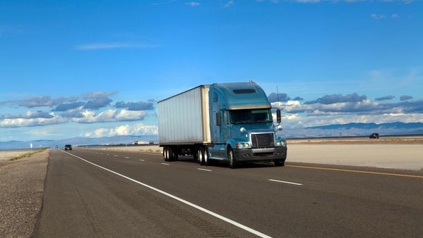 Blue semi-truck driving on a wide highway under a bright sky