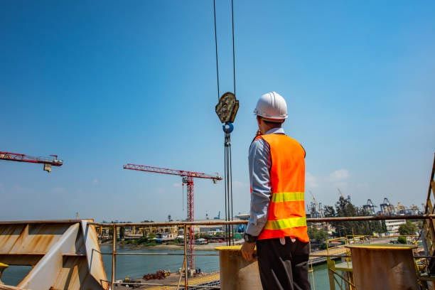 Construction worker in safety vest and hardhat looks up at crane hook.