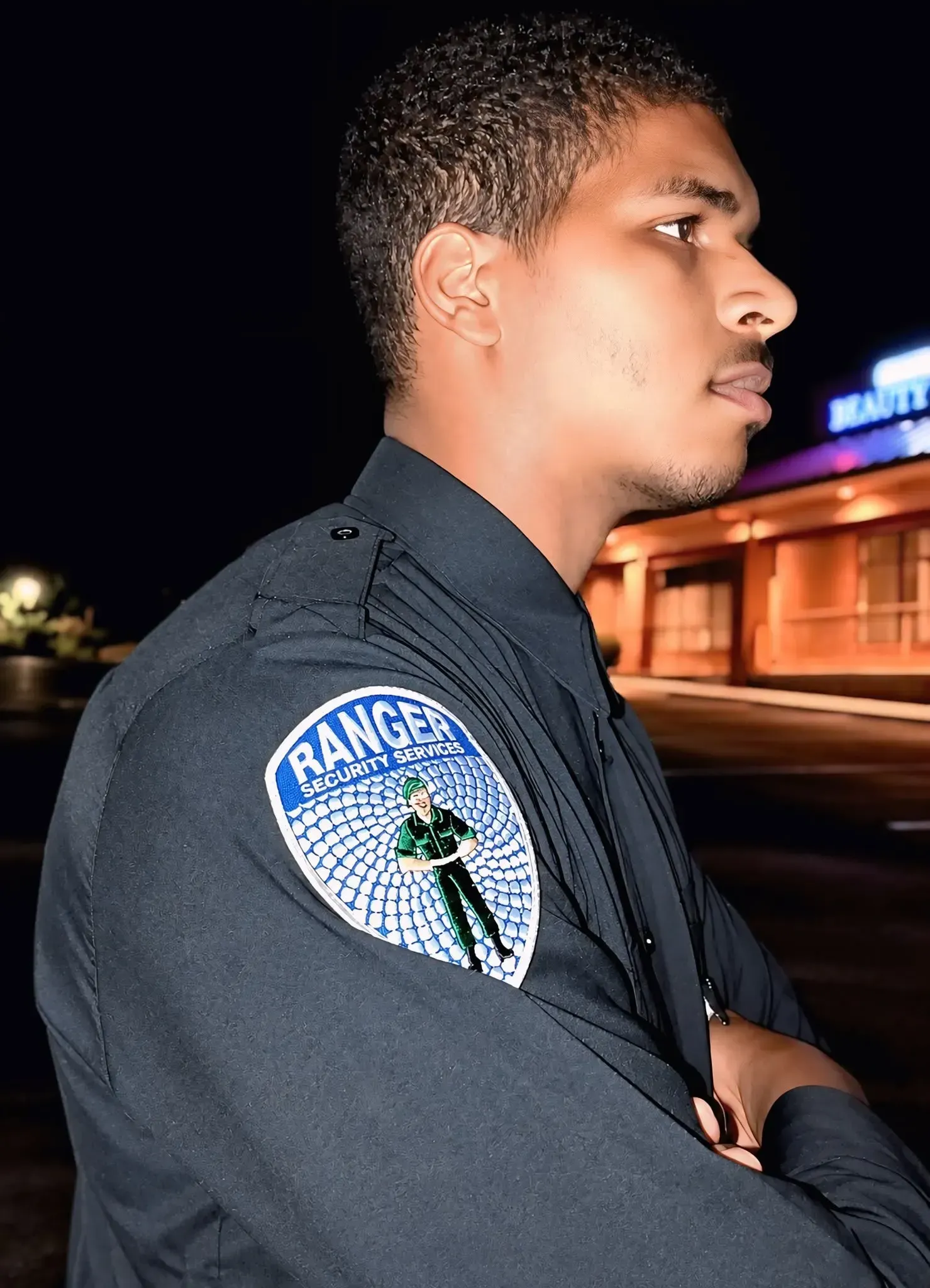 Profile of a person in a dark uniform with a badge, standing outdoors at night near a lit building.