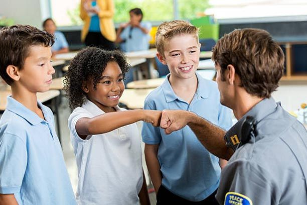 Police officer fist-bumps three students in a school classroom. One student smiles, others look on.