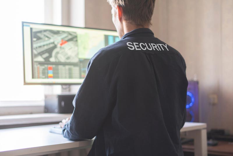 Person in security uniform monitors a computer, CCTV feeds visible on screen, white desk, and blue computer.