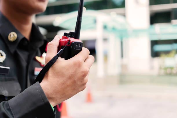 Person in dark uniform holding a red walkie-talkie outside a building.