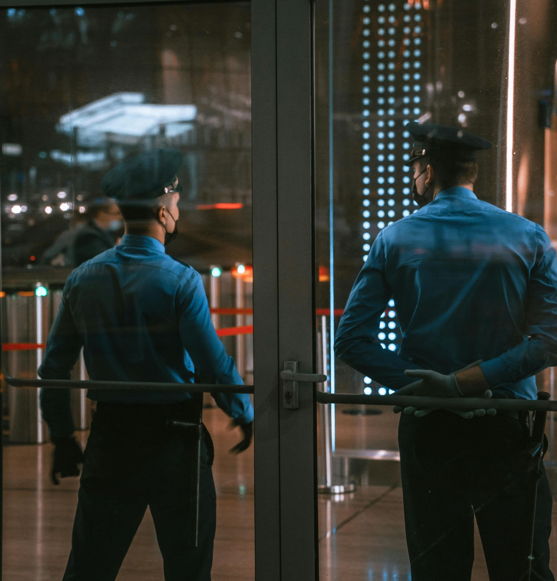 Two security guards in blue uniforms stand at a glass entrance at night