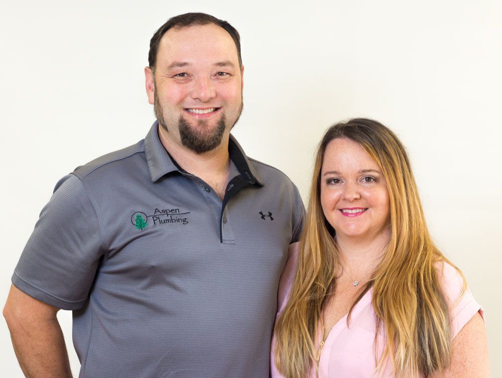 Man and woman smiling, posed in front of a neutral backdrop. Man wears a gray shirt with a logo, woman a pink top.