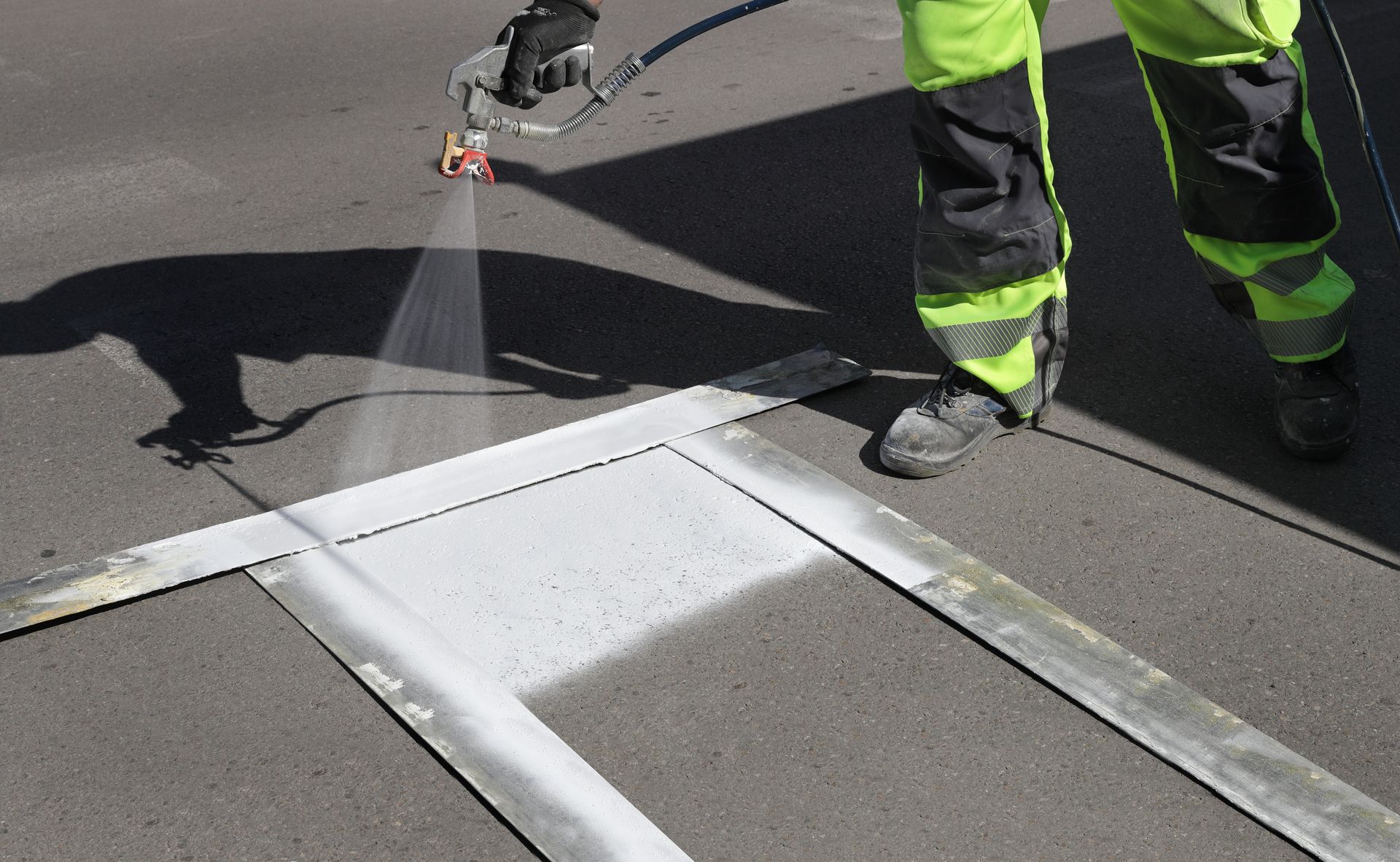 A worker in high-visibility clothing uses a spray gun to paint a white road marking using a metal stencil.