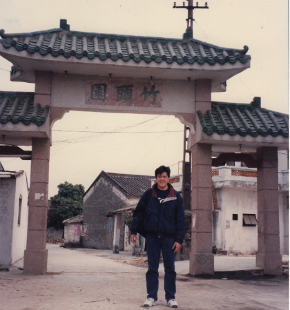 Man standing under an ornate archway in front of a Chinese village.