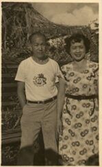 A man and woman pose outdoors. The man in a t-shirt and khakis, the woman in a floral dress.