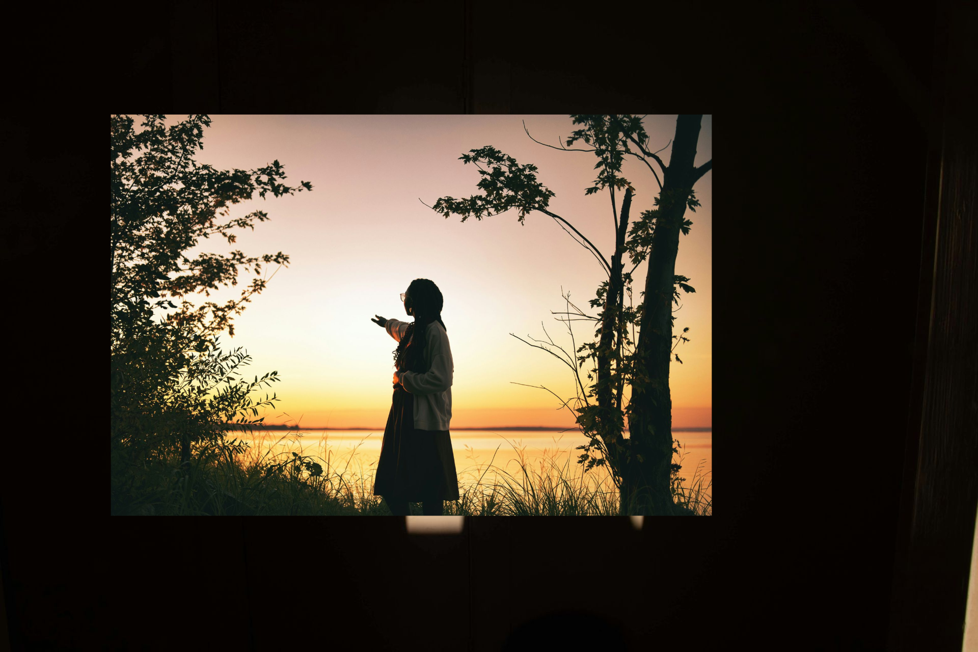 Silhouette of a person by a lakeside at sunset, framed by trees and glowing orange sky