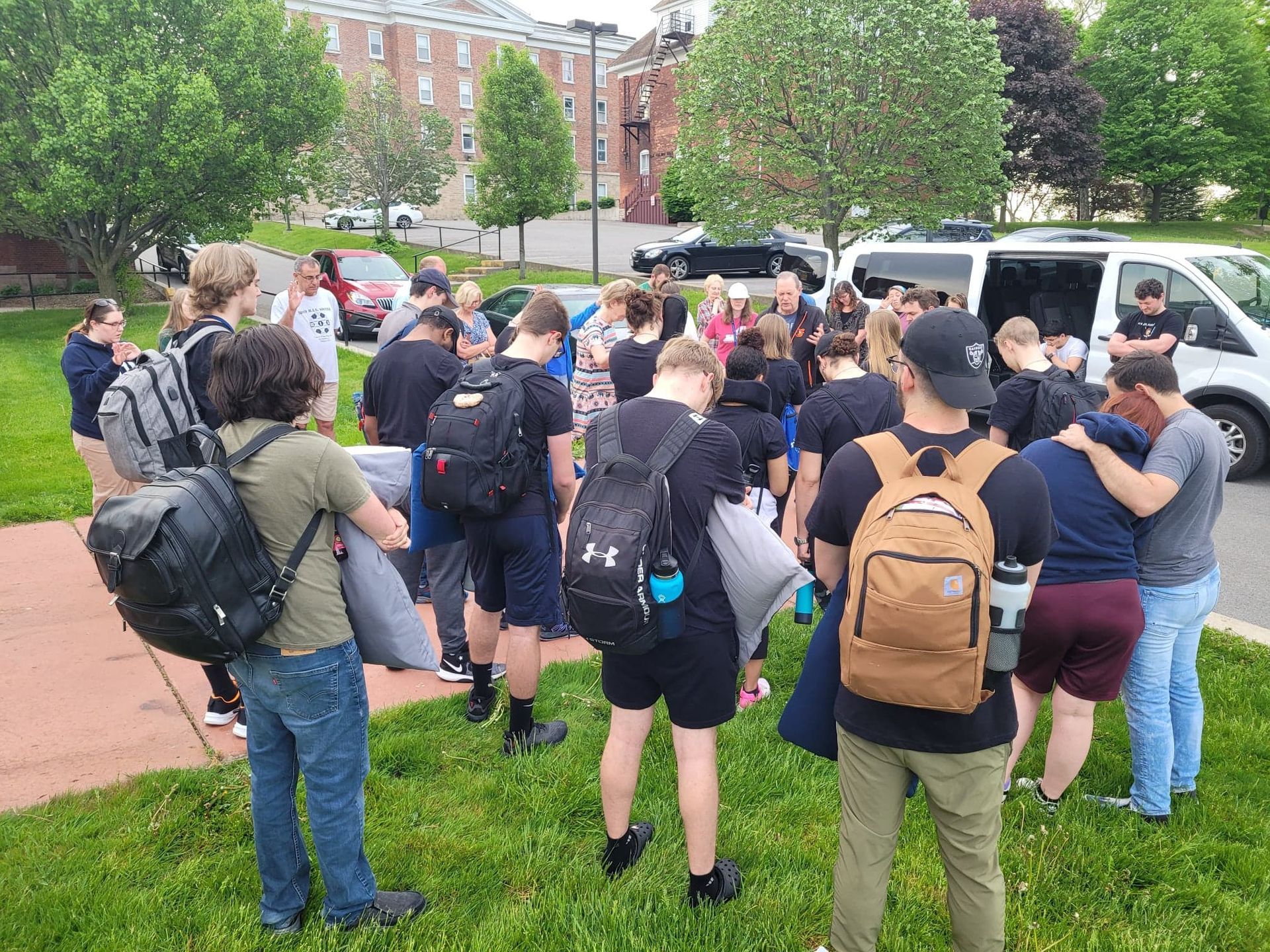 A group of young people gather outdoors, many wearing black, some holding pillows and bags. A van is parked nearby.