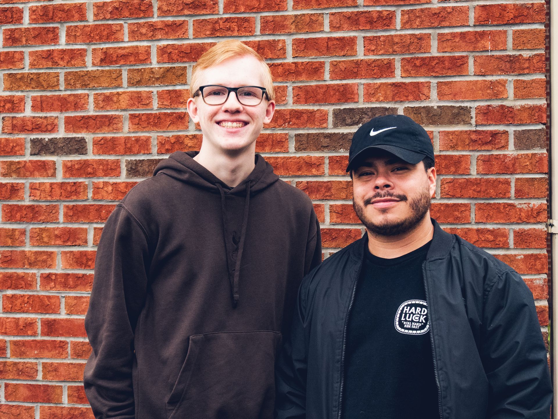 Two young men are standing next to each other in front of a brick wall.