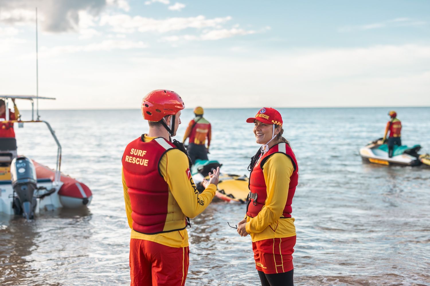 Two rescue workers in red gear by the sea, with jet skis and a boat nearby