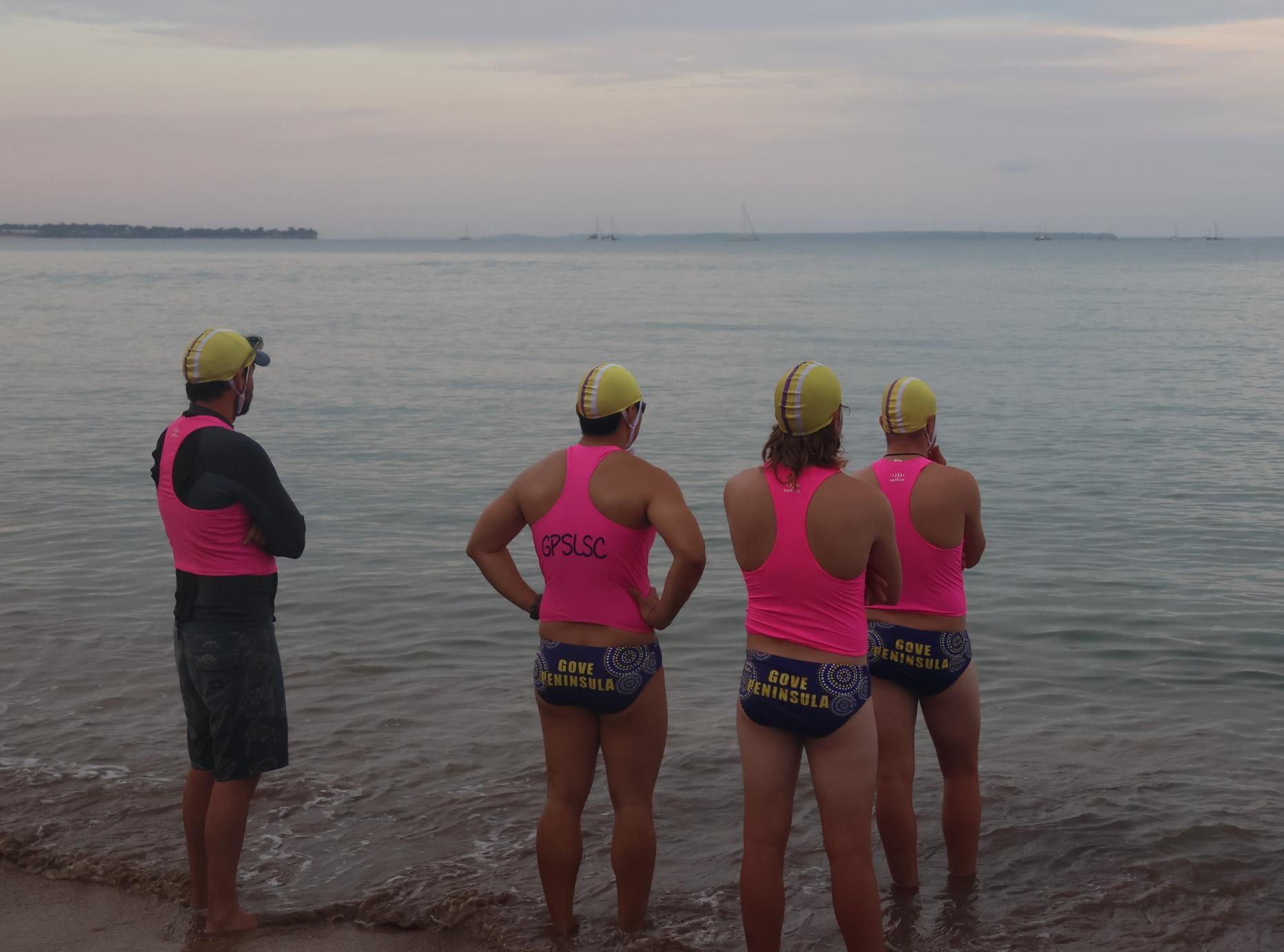 Four runners in pink tops and yellow caps stand at the shoreline facing the calm sea.