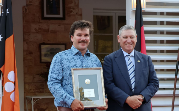 Two people holding a framed plaque at an indoor ceremony, with flags and a poster in the background