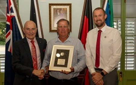Three men pose indoors, one holding a framed plaque, with flags and a framed portrait behind them.