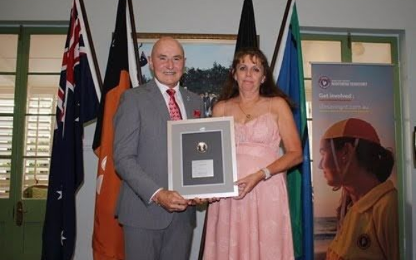 Two people holding a framed plaque at an indoor ceremony, with flags and a poster in the background