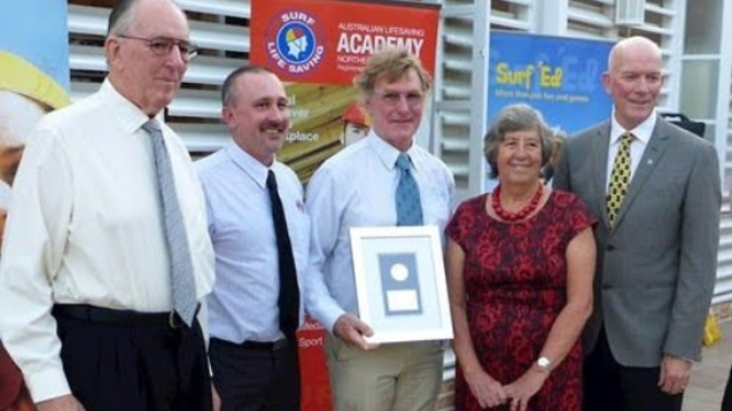 Five people posing outdoors, one holding a framed award, with an academy banner in the background