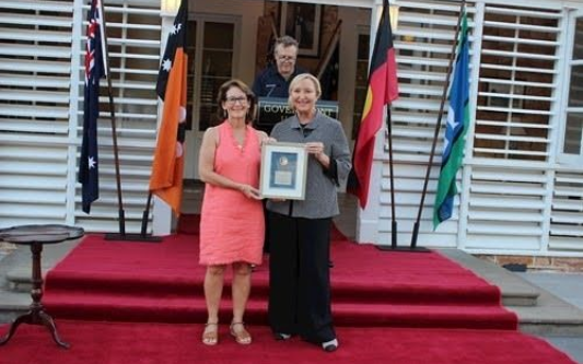 Two people on a red-carpeted stage, one holding a framed award in front of flags and window shutters.