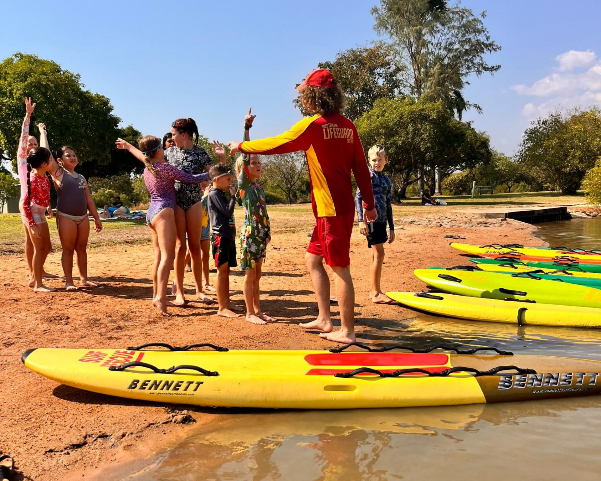 People on a riverbank holding kayaks, with yellow kayaks in the foreground and trees under a blue sky.
