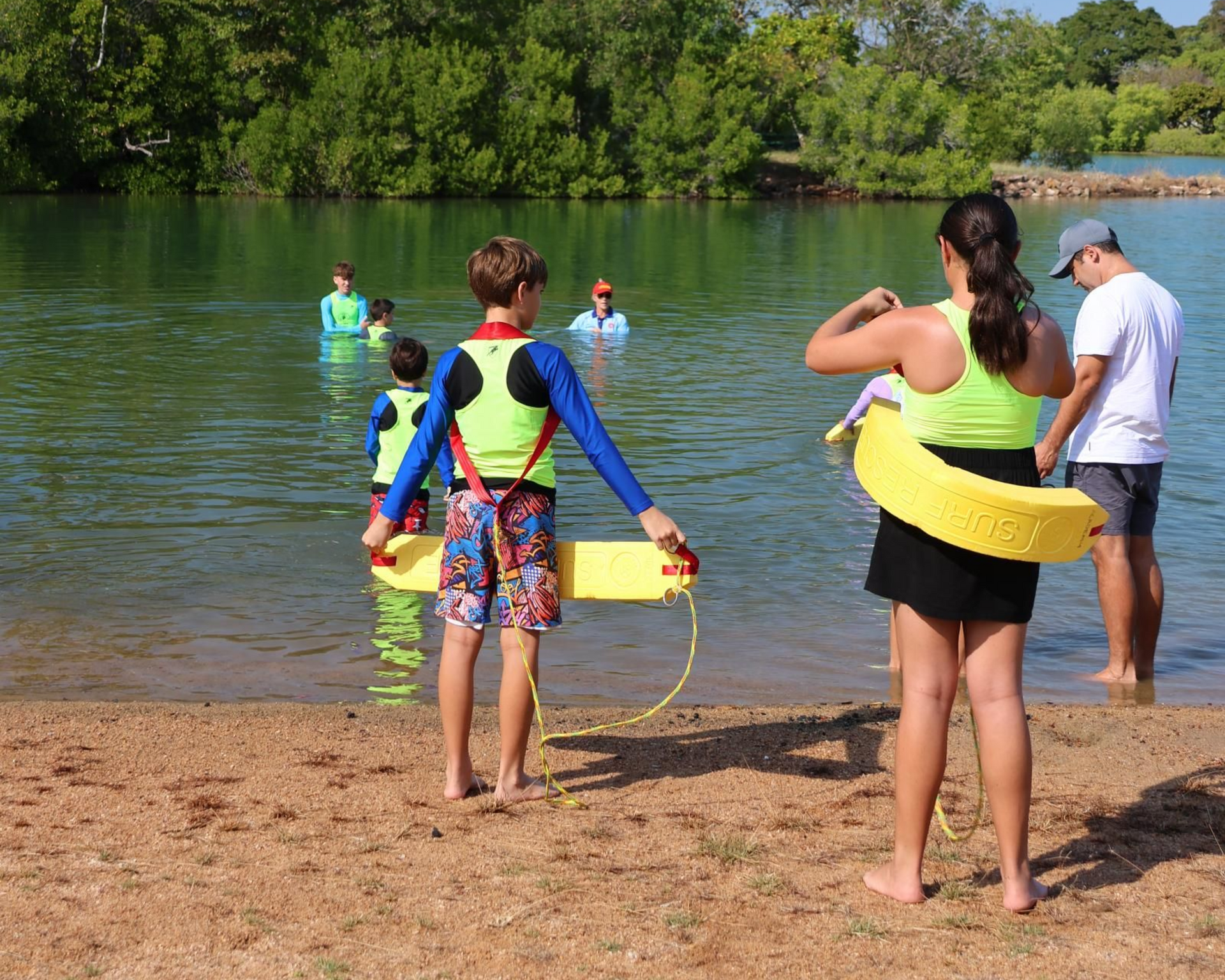 People at a lake shore wearing swim gear and flotation rings, watching swimmers in the water.
