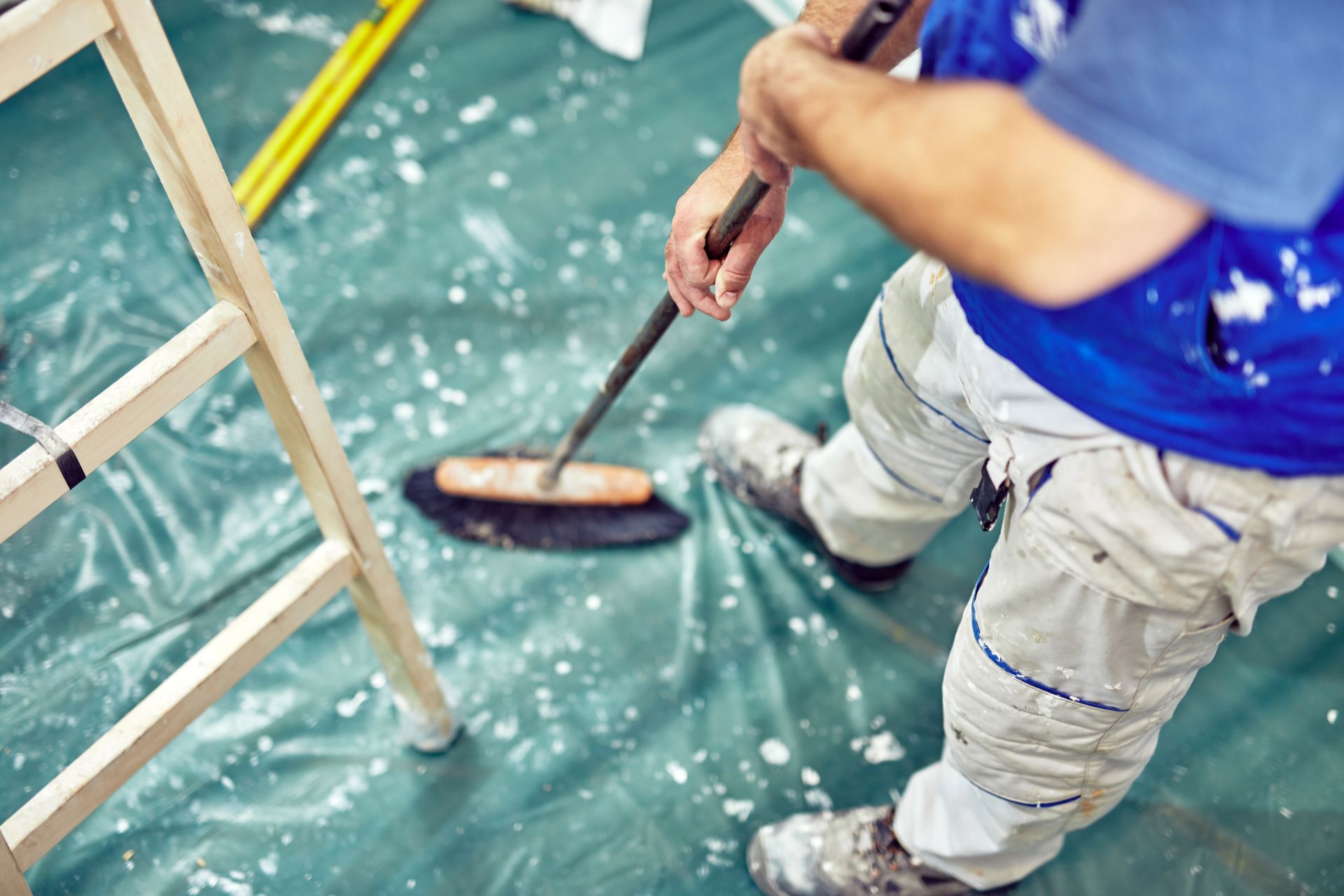 Painter using a roller on a floor, with ladder, plastic sheeting, and paint splatters.
