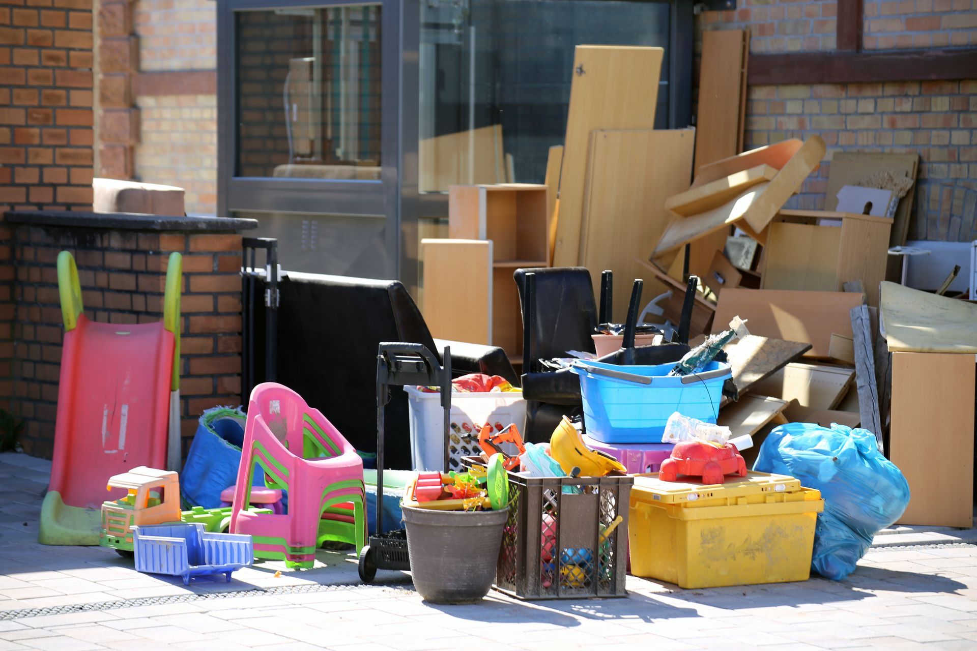 Pile of discarded items, including furniture, toys, and trash bags, on a paved surface near a building.