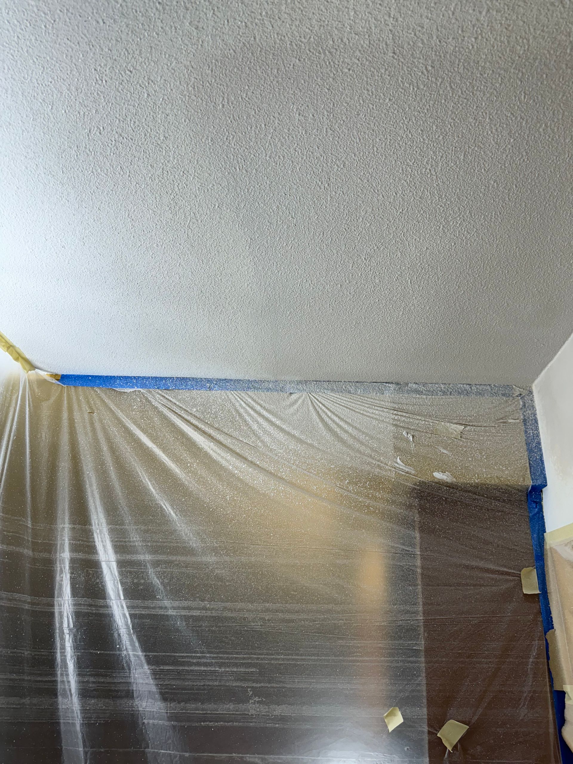 Ceiling with popcorn texture being painted; walls are covered in plastic for protection.