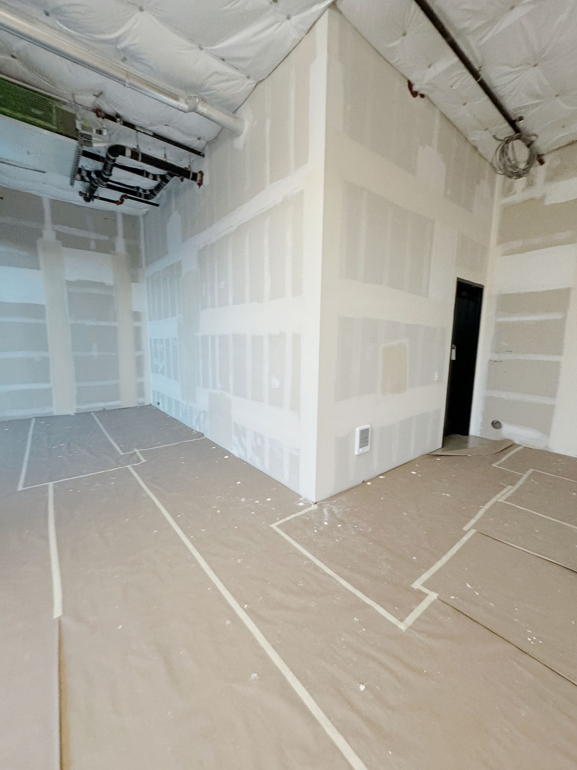 Interior view of a room under construction, with drywall on walls and floor covered with protective material.